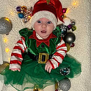 Naya a rejoint le concours — aidez-le/la à gagner de superbes lots ! baby, celebration, child, christmas, christmas_balls, cute, elf_costume, fairy_lights, festive, golden_star, green_dress, holiday, lying_down, ornaments, red_hat, striped_sleeves, surprised_expression, tights, white_blanket, winter