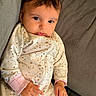 baby, child, infant, portrait, sitting, couch, gray_couch, onesie, pajamas, patterned_clothing, long_sleeves, pink_cuffs, dark_hair, brown_eyes, wide_eyed, looking_at_camera, indoors, soft_lighting, close_up, adorable