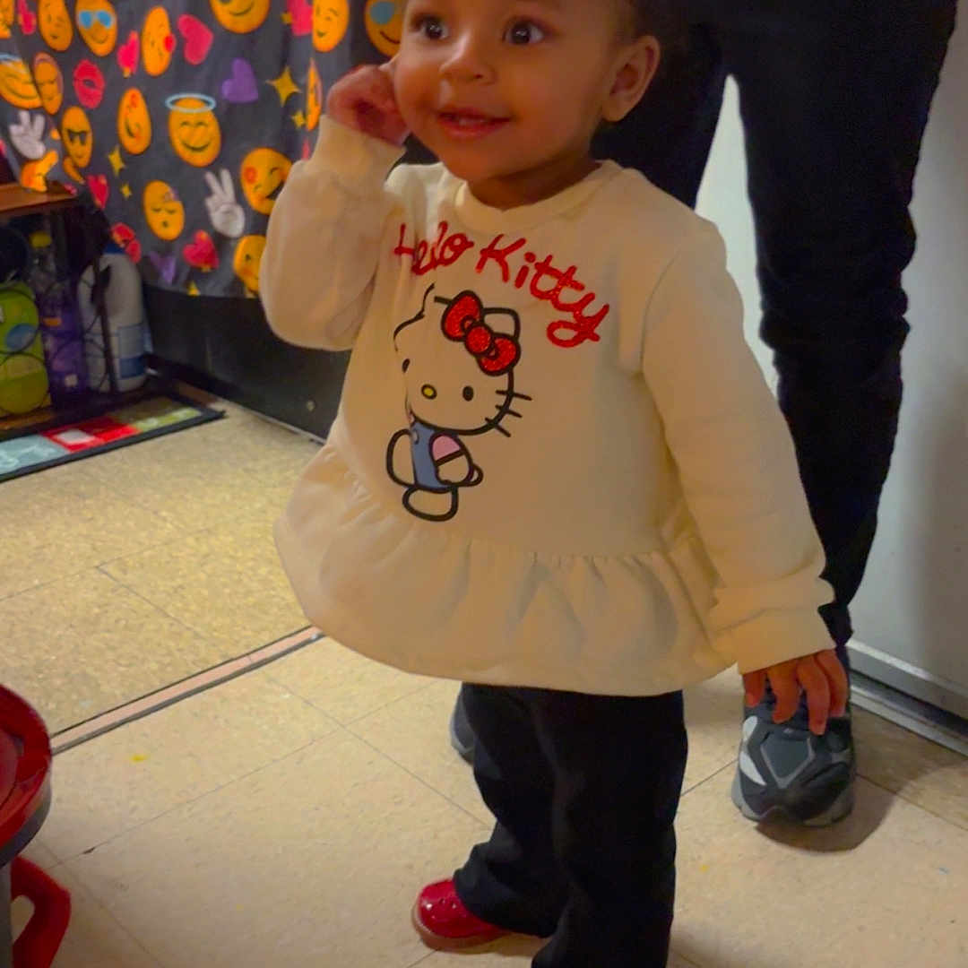 Delynn joined the competition — help win amazing prizes! toddler, child, indoor, hello_kitty, sweatshirt, red_shoes, curly_hair, person, floor, curtain, emojis, smiling, standing, clothing, adult, background, happy, hair_tie, footwear, tile_floor