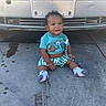 toddler, child, baby, sitting, smiling, curly_hair, blue_shirt, checkered_shorts, socks, car, license_plate, concrete, driveway, tire_marks, water_spots, toy, outdoor, daylight, front_view, person