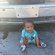 Kaida is registered to the contest to win money with this photo: baby, child, toddler, sitting, concrete, car, license_plate, blue_shirt, checkered_shorts, socks, pacifier, curly_hair, outdoor, daylight, vehicle, grill, head, face, smile, clothing