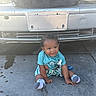 baby, child, toddler, sitting, concrete, car, license_plate, blue_shirt, checkered_shorts, socks, pacifier, curly_hair, outdoor, daylight, vehicle, grill, head, face, smile, clothing