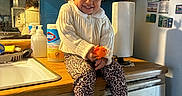 Eliana is registered to the contest to win money with this photo: child, toddler, smiling, sitting, toy, kitchen, counter, paper_towel, cleaning_supplies, sink, cabinet, curly_hair, bow, leopard_print, slippers, wall_notes, reflection, indoor, clothing, happy