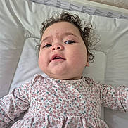 Mia participe au concours pour gagner de l'argent avec cette photo : baby, child, curly_hair, floral_dress, cushion, white_background, face, skin, expression, lying_down, infant, portrait, indoors, cute, person, head, clothing, young_child, health, closeup