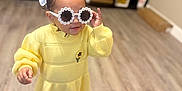 Josemid is registered to the contest to win money with this photo: toddler, child, indoor, floor, yellow_dress, sunglasses, hair_bow, boots, person, smiling, cute, fashion, standing, wood_floor, furniture, shelf, home, playful, portrait, accessory