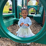 Josemid is registered to the contest to win money with this photo: child, toddler, playground, play_structure, green, circular_frame, smiling, outdoor, daylight, woodchips, skirt, sweater, hair_accessories, shoes, happy, person, nature, grass, park, trees