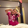 bed, bedroom, blanket, blurred_background, casual_clothing, child, cozy, furniture, girl, hand_gesture, indoor, long_hair, photograph, pink_shirt, playful, portrait, sitting, sleeping_area, smiling, warm_lighting