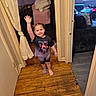 air_conditioner, bedroom, braided_hair, child, couch, doorframe, girl, hallway, home, indoor, mismatched_socks, pigtails, pillow, shorts, smiling, toy_car, tshirt, waving, window, wooden_floor