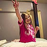 child, girl, pink_shirt, bed, blanket, smile, peace_sign, long_hair, indoor, mirror, hat, curtain, wall, person, casual_clothing, home, portrait, happy, gesture, room