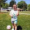 bandage, casual_clothing, child, daytime, fun, grass, happy, heart_shaped, outdoor, park, person, playground, shoes, shorts, smiling, soccer_ball, sunglasses, sunny, tree, tshirt