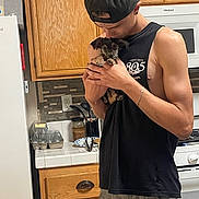 Hiccup joined the competition — help win amazing prizes! man, puppy, dog, kitchen, holding, cap, black_shirt, wooden_cabinets, stove, refrigerator, countertop, affection, pet, casual_clothing, home_interior, young_adult, small_dog, appliance, indoor, love