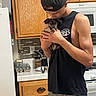 man, puppy, dog, kitchen, holding, cap, black_shirt, wooden_cabinets, stove, refrigerator, countertop, affection, pet, casual_clothing, home_interior, young_adult, small_dog, appliance, indoor, love