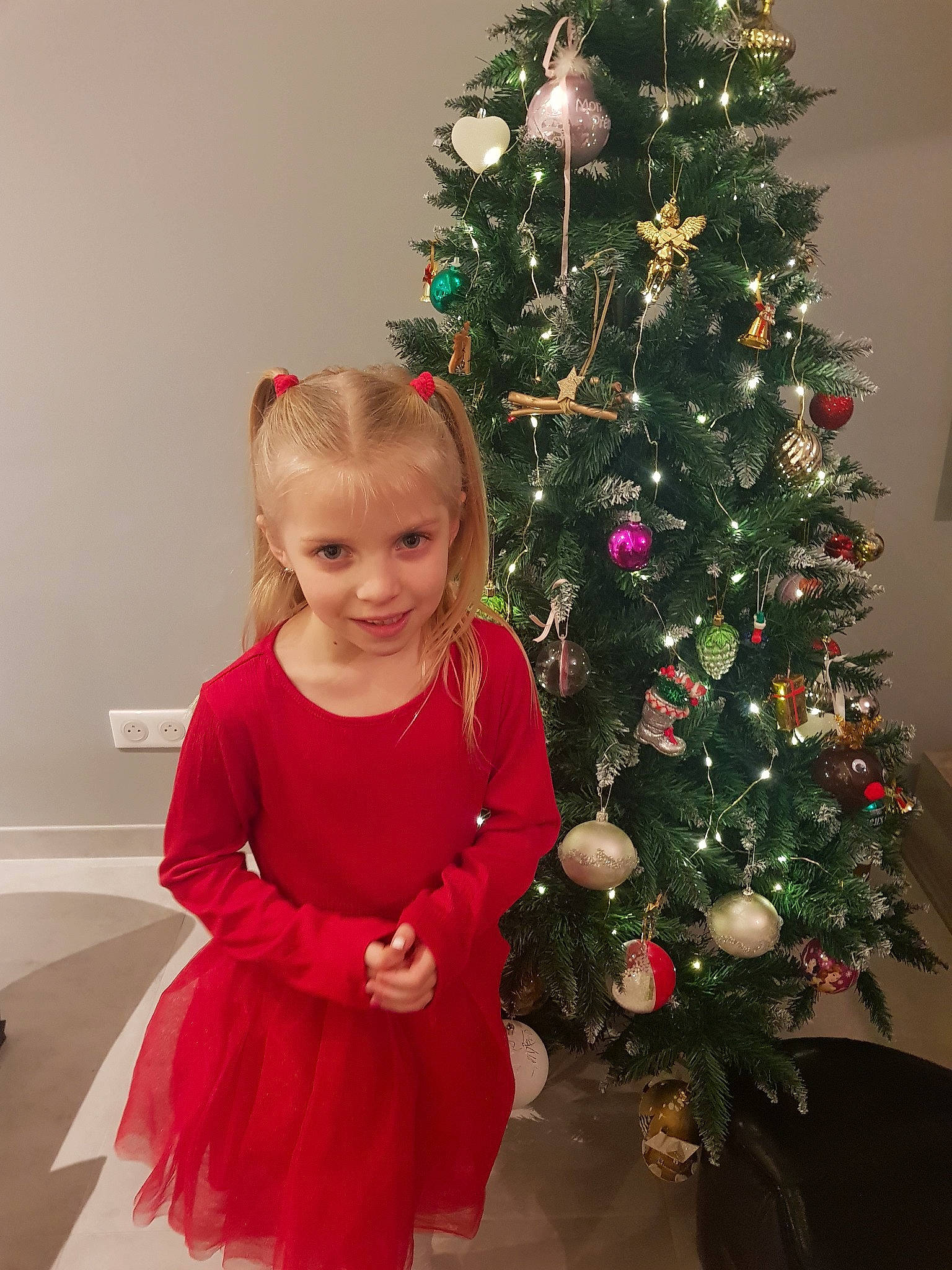 Emma participe au concours pour gagner de l'argent avec cette photo : baby_toddler_clothing, christmas_decoration, christmas_ornament, christmas_tree, dress, event, evergreen, facial_expression, happy, head, holiday, holiday_ornament, human_body, joy, ornament, person, plant, smile, standing, tints_and_shades