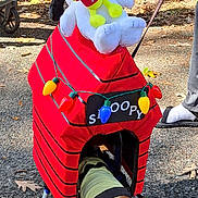Sammy is registered to the contest to win money with this photo: dog, wheelchair, costume, snoopy, woodstock, plush_toy, christmas_lights, red, yellow, outdoor, pavement, leaves, tree, person, footwear, sunlight, shadow, pet, animal, cute
