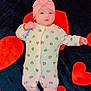 baby, infant, onesie, pink_hat, turban, lying_down, blanket, heart_pattern, red_hearts, black_blanket, cute, portrait, child, sleepwear, footed_pajamas, soft_texture, indoor, adorable, face, young_child