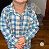 child, toddler, smiling, plaid_shirt, standing, indoor, wooden_floor, hands_clasped, happy, person, casual_clothing, green_shoes, brown_hair, cute, portrait, home, flooring, young_child, cheerful, bright_lighting