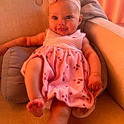 Scarlett is registered to the contest to win money with this photo: baby, child, infant, pink_dress, headband, bow, couch, pillow, smile, face, legs, arm, foot, indoor, cute, happy, person, furniture, portrait, relaxing