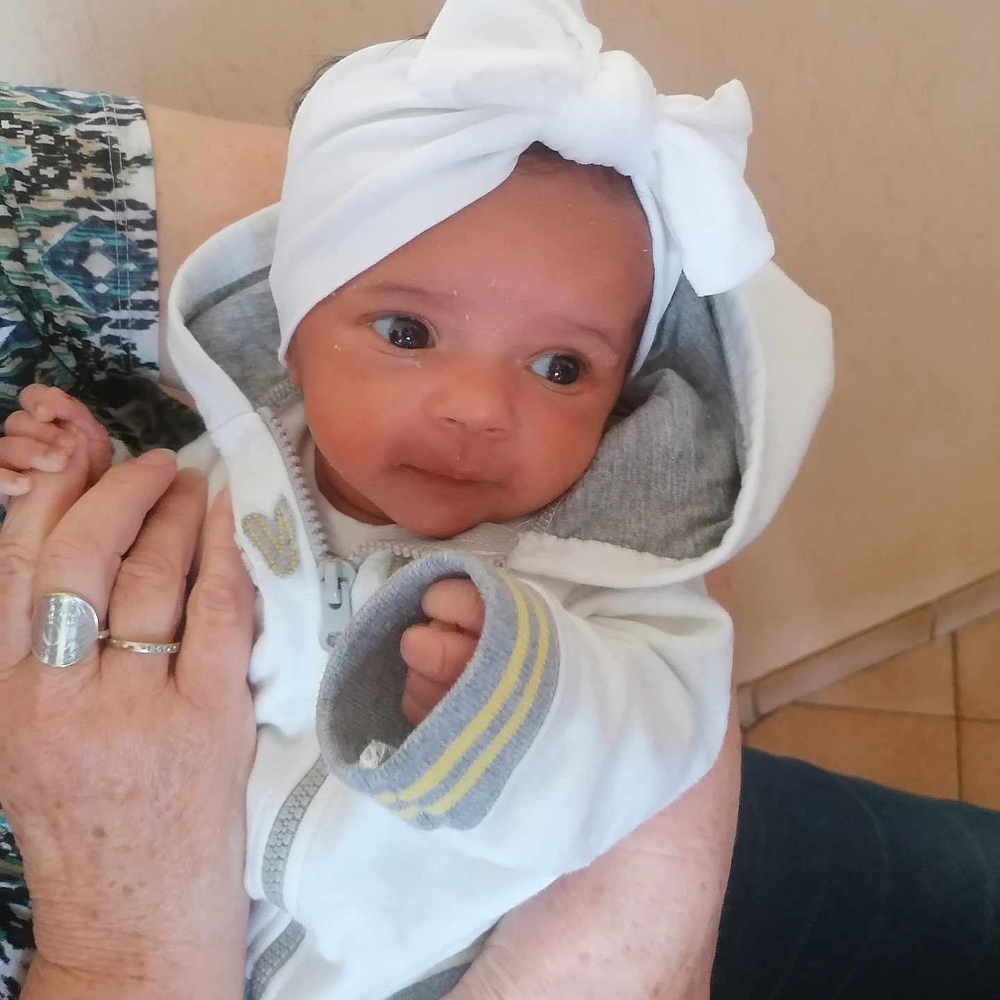 Luna a rejoint le concours — aidez-le/la à gagner de superbes lots ! adult_hand, baby, bow, child, closeup, cozy, cute, expression, face, hand, headband, holding, indoor, infant, patterned_clothing, person, portrait, rings, warm_clothing, white_hoodie