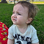 Xavier is registered to the contest to win money with this photo: toddler, child, grass, outdoor, mickey_mouse, shirt, red_blanket, cartoon, expression, confused, displeased, sunny, daylight, park, tree, person, casual_clothing, young_child, nature, sitting