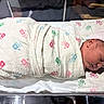 newborn, baby, infant, sleeping, swaddled, blanket, cap, hospital, bassinet, portrait, face, closed_eyes, peaceful, cozy, handprint_pattern, pink, white, nursery, lying_down, child