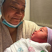 Osana is registered to the contest to win money with this photo: adult, baby, newborn, mother, parent, hospital, portrait, close_up, face, smiling, looking_down, infant, blanket, swaddle, knit_cap, medical_mask, clothing, caregiving, indoor, family
