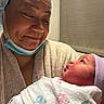 adult, baby, newborn, mother, parent, hospital, portrait, close_up, face, smiling, looking_down, infant, blanket, swaddle, knit_cap, medical_mask, clothing, caregiving, indoor, family