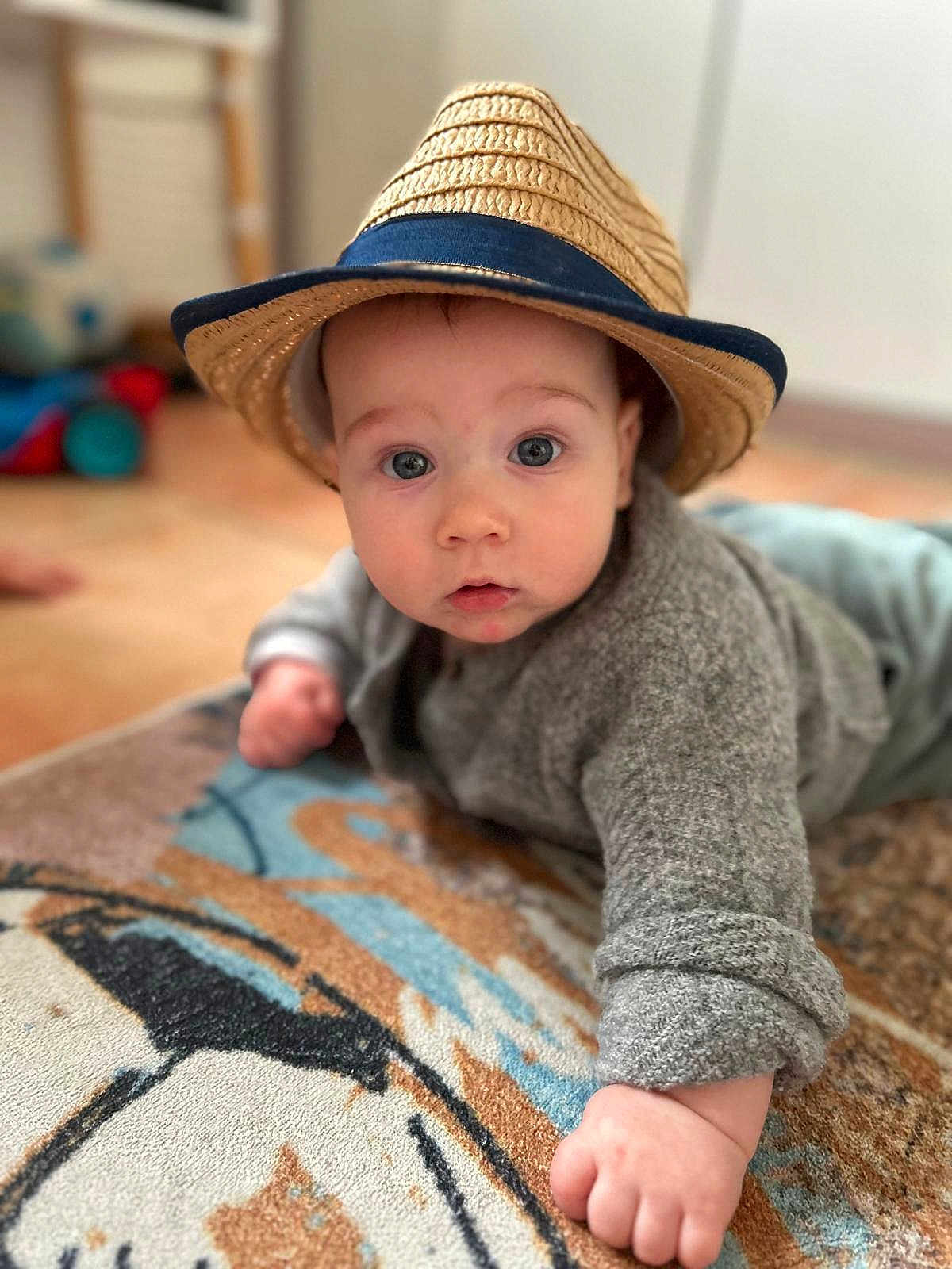 Julian a rejoint le concours — aidez-le/la à gagner de superbes lots ! baby, child, hat, straw_hat, gray_sweater, rug, floor, indoor, portrait, face, hand, curious, wide_eyes, cozy, cute, infant, crawling, patterned_rug, soft_focus, expression