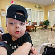 Ryder is registered to the contest to win money with this photo: toddler, child, cap, black_clothing, indoor, waiting_room, chairs, plant, tile_floor, person, hand, adult, curious, holding, face, portrait, casual, baby, hat, room