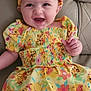baby, child, smiling, dress, headband, yellow, cartoon_print, sitting, couch, leather, happy, portrait, indoor, cute, infant, person, clothing, colorful, face, young