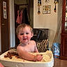 Oaklee is registered to the contest to win money with this photo: toddler, child, high_chair, food, indoors, kitchen, wall_decor, sunflower, wooden_furniture, person, baby, smiling, face, hair, tray, seat, floor, door, coat, cabinet