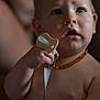 baby, infant, pacifier, blue_eyes, necklace, skin, hand, face, expression, curious, child, portrait, closeup, indoors, soft_lighting, warm_tones, blurry_background, holding, mouth_open, caucasian