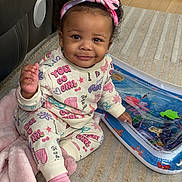 Navaya is registered to the contest to win money with this photo: toddler, child, smiling, headband, colorful_clothing, pink_socks, blanket, water_mat, carpet, indoor, cute, happy, sitting, playful, floor, person, young_child, curly_hair, cozy, home