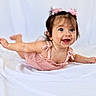 baby, child, infant, smiling, happy, portrait, pink_outfit, dress, hair_bows, flower_clips, lying_down, arms_outstretched, indoor, white_background, bedding, blanket, baby_photography, close_up, joyful, playful