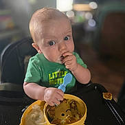 Richard is registered to the contest to win money with this photo: baby, child, high_chair, green_shirt, spoon, food, snack, plate, blue_spoon, face, eyes, hand, meal, kitchen, indoor, curious, eating, messy, person, toddlers