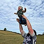 baby, adult, child, outdoor, field, grass, sky, clouds, person, holding, playing, hat, vest, jeans, smiling, nature, happy, daylight, fun, family