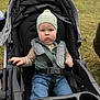 baby, child, stroller, hat, vest, jeans, outdoor, grass, parked_cars, straps, serious_face, person, clothing, seat, hand, wheelchair, autumn, cute, portrait, daytime