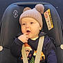 baby, child, car_seat, hat, pom_pom, finger_in_mouth, colorful_clothing, leggings, animal_print, navy_top, sitting, safety_harness, cute, indoor, window, soft_lighting, infant, expression, person, portrait