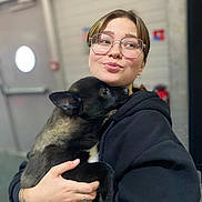 Millow a rejoint le concours — aidez-le/la à gagner de superbes lots ! person, dog, puppy, glasses, black_hoodie, indoor, hug, affection, cute, pet, young_adult, smile, short_hair, holding, animal, face, warmth, comfort, casual_clothing, love