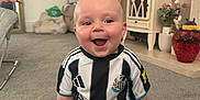 Hunter is registered to the contest to win money with this photo: baby, child, smile, sitting, carpet, soccer_jersey, black_and_white, socks, living_room, furniture, flowers, blanket, toy, happy, indoor, person, head, couch, decor, home