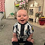baby, child, smile, sitting, carpet, soccer_jersey, black_and_white, socks, living_room, furniture, flowers, blanket, toy, happy, indoor, person, head, couch, decor, home