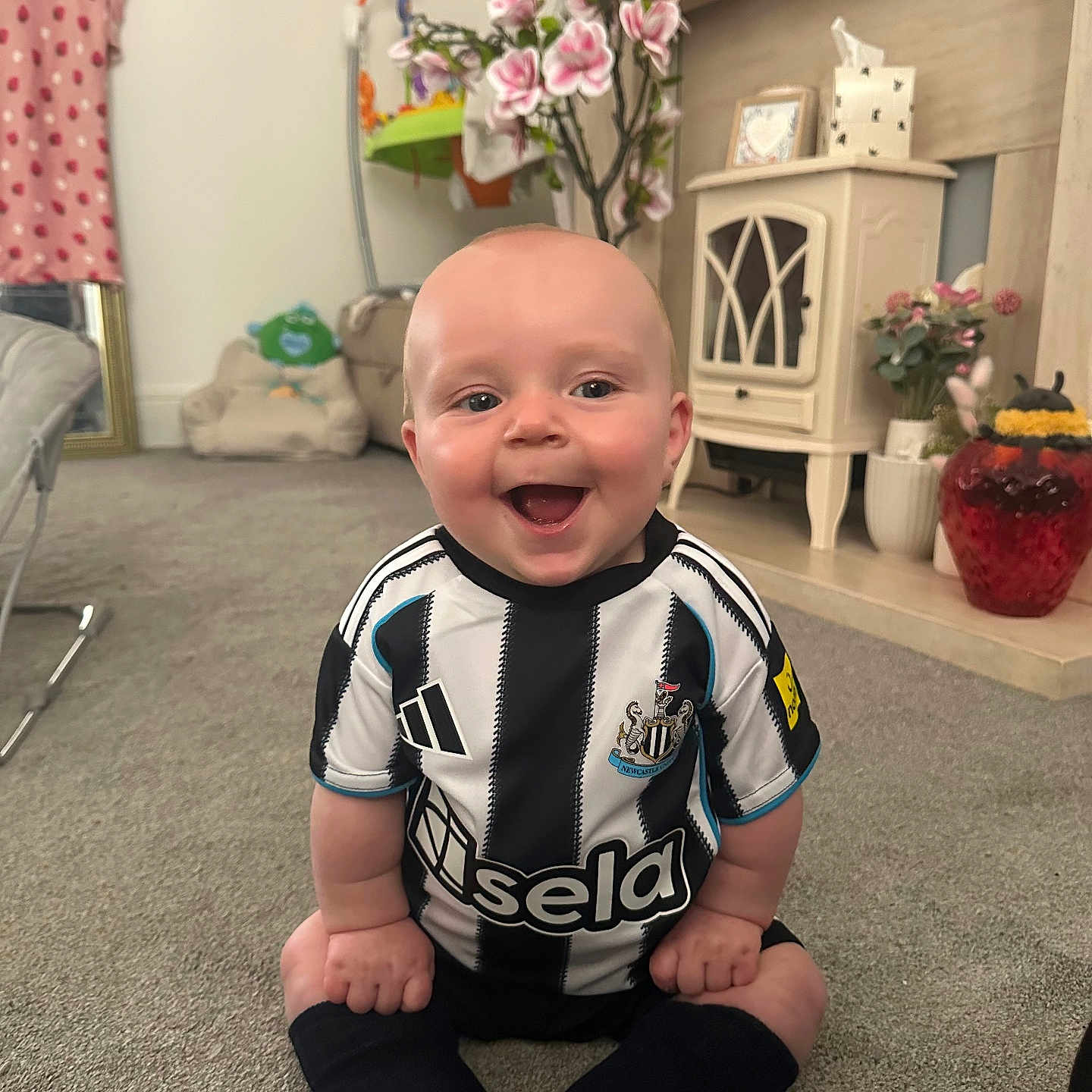 Hunter is registered to the contest to win money with this photo: baby, black_and_white, blanket, carpet, child, couch, decor, flowers, furniture, happy, head, home, indoor, living_room, person, sitting, smile, soccer_jersey, socks, toy