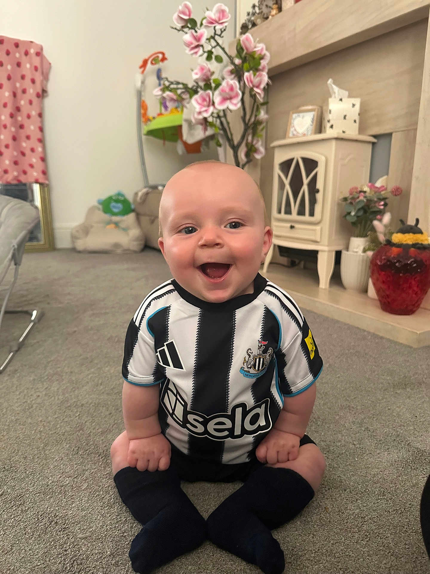 Hunter is registered to the contest to win money with this photo: baby, child, smile, sitting, carpet, soccer_jersey, black_and_white, socks, living_room, furniture, flowers, blanket, toy, happy, indoor, person, head, couch, decor, home