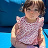 toddler, child, baby, curly_hair, blue_cushion, floral_dress, pink_socks, sitting, outdoor, sunlight, portrait, cute, innocent, expression, person, face, young_child, casual_clothing, relaxed, closeup