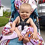 baby, blanket, car, child, clothing, face, feet, grass, hand, happy, holding, outdoor, parking_lot, person, purple, sidewalk, smiling, stroller, toy, vehicle