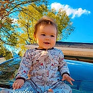 Taylor is registered to the contest to win money with this photo: baby, child, pajamas, car, hood, outdoor, sky, trees, sunlight, smile, happy, nature, portrait, infant, clothing, blue_sky, greenery, time_stamp, sitting, cute