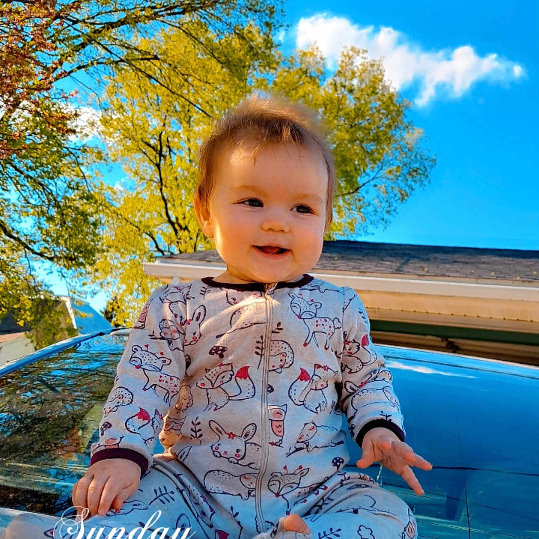 Taylor is registered to the contest to win money with this photo: baby, child, pajamas, car, hood, outdoor, sky, trees, sunlight, smile, happy, nature, portrait, infant, clothing, blue_sky, greenery, time_stamp, sitting, cute