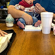 Melesia is registered to the contest to win money with this photo: baby, adult, table, wooden_table, bottle, cup, book, reading, clothing, denim, sitting, hands, expression, indoor, person, face, holding, casual, surprised, focus