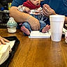baby, adult, table, wooden_table, bottle, cup, book, reading, clothing, denim, sitting, hands, expression, indoor, person, face, holding, casual, surprised, focus