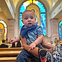 baby, child, indoor, church, stained_glass, colorful, holding, person, portrait, curious, blue_clothing, scarf, hands, bench, religious, architecture, wristband, patterned_clothing, seated, people