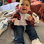 Paxton is registered to the contest to win money with this photo: baby, child, smiling, jeans, shirt, white_shoes, blanket, bed, indoors, person, happy, clothing, plaid, sitting, cute, holding, toy, casual, footwear, portrait
